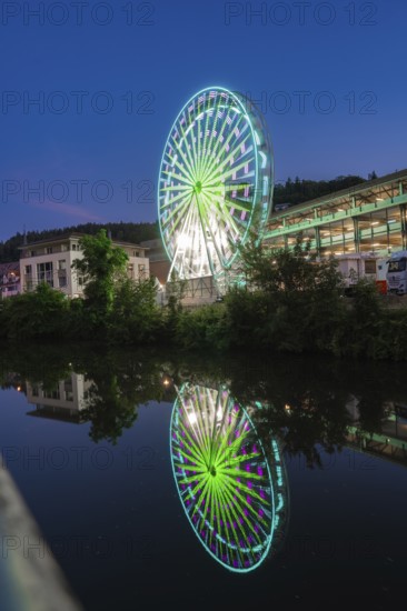A red illuminated Ferris wheel with reflection in the river against a city backdrop in the evening, 950 years of Calw, Germany