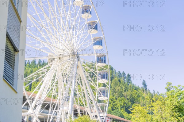A large Ferris wheel stands next to a building with a wooded hill and clear sky in the background, 950 years of Calw, Germany