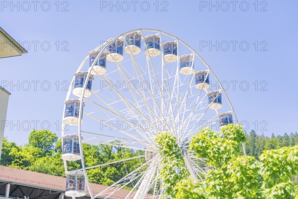 A white Ferris wheel turns in front of a building surrounded by green vegetation on a clear summer day, 950 years Calw, Germany
