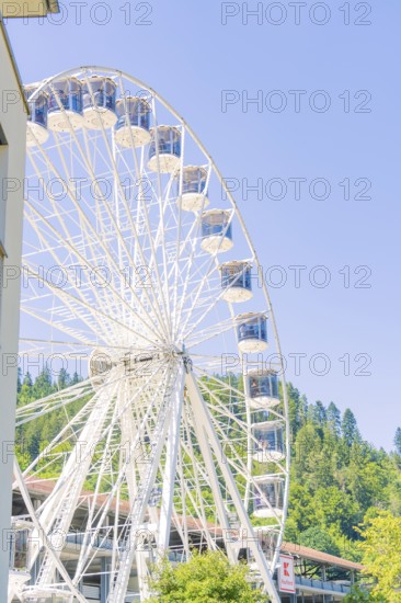 A Ferris wheel with numerous cabins stands close to a building, against a green background and blue sky, 950 years Calw, Germany