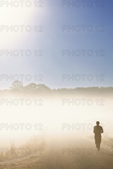 Man running on a dirt road in morning fog in the countryside with the sun shining through the fog