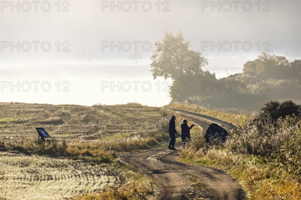 Couple standing on a winding gravel road in a rolling agricultural landscape with low morning mist on the fields looking on the view a beautiful sunny autumn morning