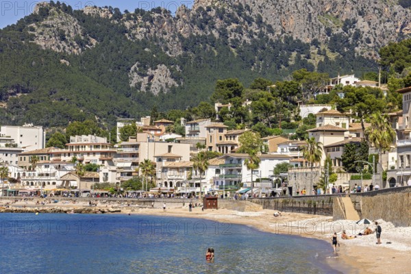 People sunbathing and bathing on the seashore at a idyllic coast village with houses at the boardwalk by the seabay and high mountains a sunny summer day, Port de Soller, Mallorca, Spain
