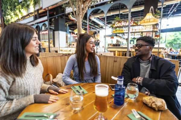 Three friends enjoying drinks and engaging in lively conversation at a stylish wooden table in a bustling modern food hall