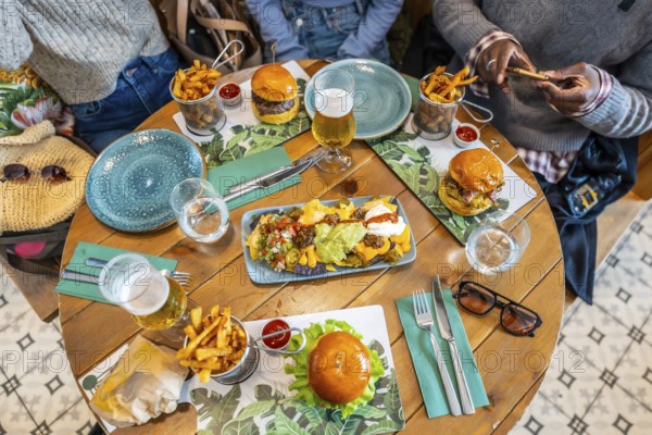 Overhead view of friends sharing a meal of burgers, nachos, french fries, and drinks at a wooden restaurant table