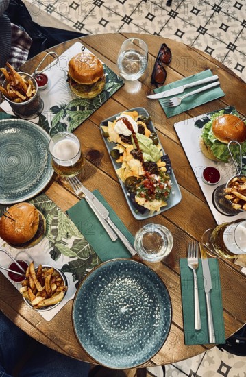Top view of a wooden restaurant table with nachos, burgers, french fries, and beer, suggesting a shared meal among friends