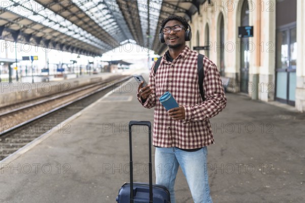 Young black male tourist waiting for his train while listening to music and holding a reusable water bottle, promoting sustainable travel