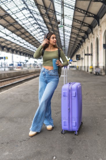 Stylish tourist listening to music while waiting for her train at a station platform, enjoying a coffee from her reusable cup
