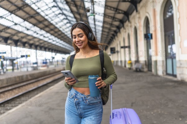 Young woman tourist walking in train station using smartphone and headphones while carrying reusable coffee cup and rolling suitcase