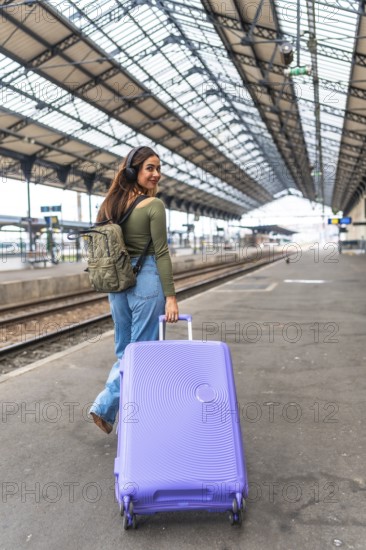 Tourist wearing headphones walking in train station pulling a purple suitcase and carrying a backpack