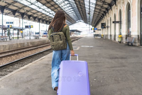 Tourist walking on railway platform pulling trolley suitcase and wearing backpack, traveling by train