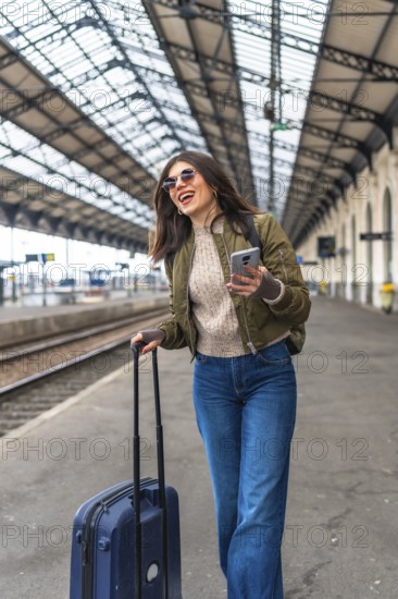 Happy female tourist walking through a bustling train station, using her smartphone while pulling a suitcase behind her