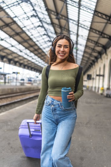 Tourist walking in train station pulling suitcase and listening to music with headphones, holding reusable coffee cup