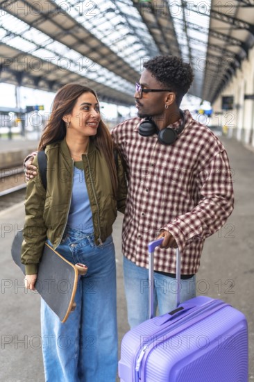 Happy multi ethnic couple walking together on platform while waiting for train, embracing and carrying luggage and skateboard