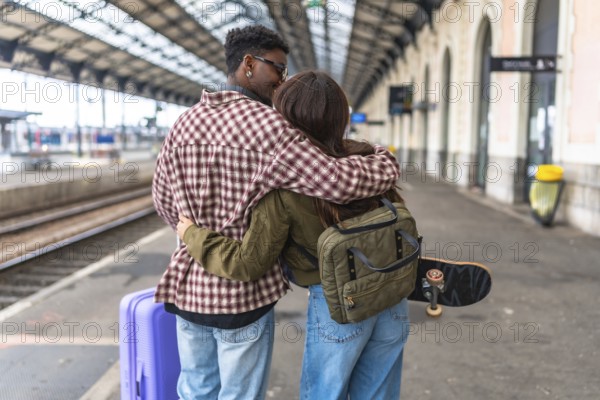 Young couple kissing and hugging on a train station platform, ready for their journey, with luggage and skateboard