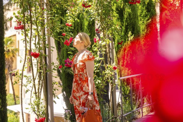 Woman in red dress smelling flowers on a plant path, surrounded by summer light, Botanical Garden, The Gardens of Trauttmansdorff Castle, Merano, South Tyrol, Italy
