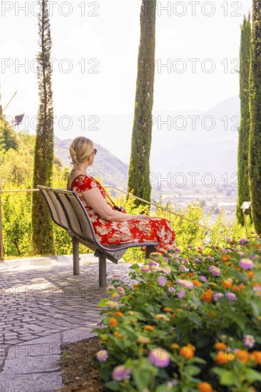 Woman in a red dress sitting on a bench surrounded by flowers and cypresses, Botanical Garden, The Gardens of Trauttmansdorff Castle, Merano, South Tyrol, Italy