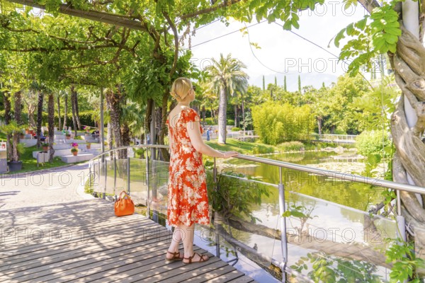 Woman in floral dress enjoying the view of a park-like pond, Botanical Gardens, The Gardens of Trauttmansdorff Castle, Merano, South Tyrol, Italy