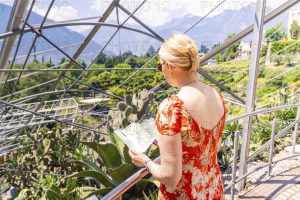 Woman in a red summer dress looking at a map in front of a glass construction, Botanical Garden, The Gardens of Trauttmansdorff Castle, Merano, South Tyrol, Italy