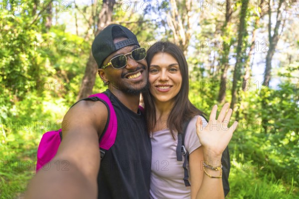 Interracial couple smiling and waving while taking a selfie during a vibrant hike in a lush, green forest filled with sunlight