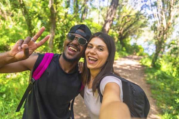 Interracial couple wearing backpacks taking a selfie while hiking in a lush forest, enjoying their outdoor adventure