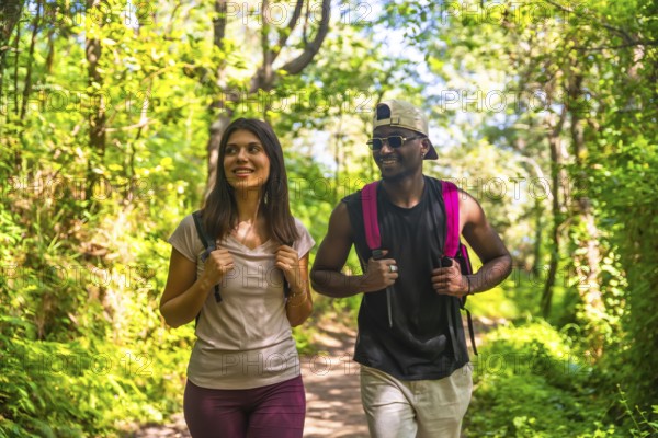 Interracial couple wearing backpacks walking in a forest trail, enjoying the beautiful nature surrounding them