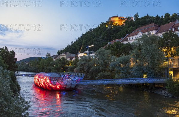 Night-time view of the Murinsel floating island on the river Mur with Schlossberg castle hill in the background, Graz, Austria
