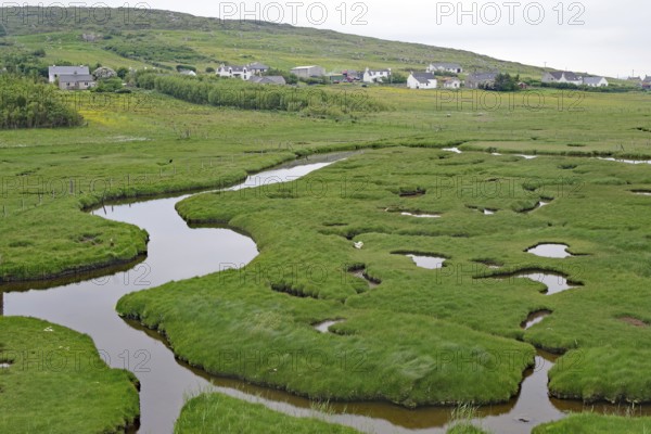 Green marshland with waterways and hills, behind a village under overcast skies, tidal landscape, cycle tour, Harris Hebrides, Scotland, Great Britain