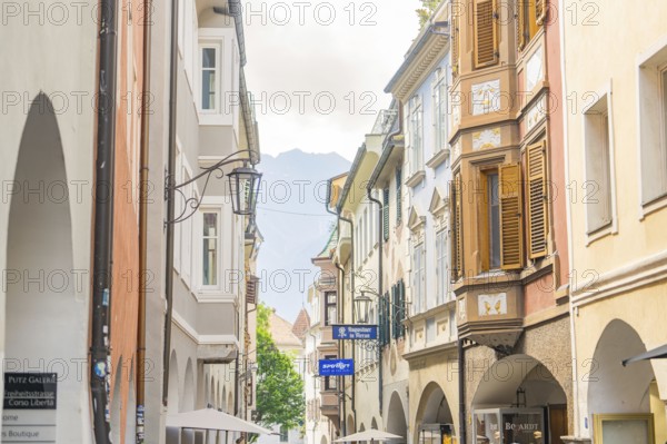 Narrow alley in the old town centre with classic buildings and shutters, mountains in the background, Merano, Italy