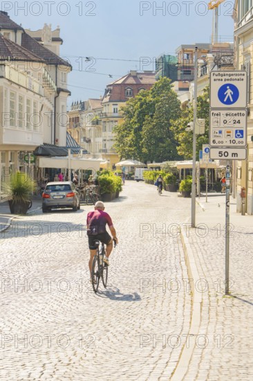 A cyclist rides along a sunny city street surrounded by historic buildings and trees, Merano, Italy