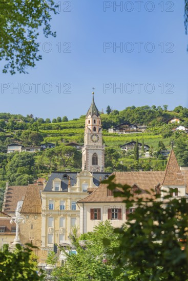 City skyline with a striking church tower surrounded by green hills against a clear sky, Merano, Italy