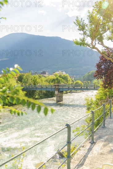 River with bridge, surrounded by green nature and mountains in sunlight, Merano, Italy