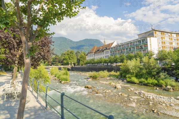 A calm river flows along an urban promenade with mountains in the background, Merano, Italy