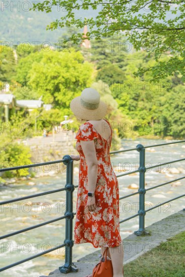 A woman in a flowered dress looks at a flowing river on a sunny day, Merano, Italy