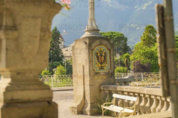 Waterfront promenade with bench and artistic coat of arms in sunny weather, Merano, Italy