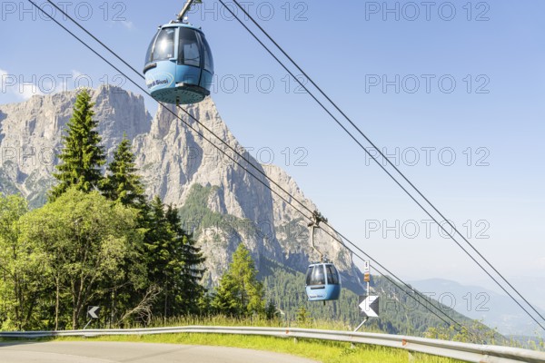 Two blue cable car gondolas hovering over trees in front of a rocky mountain range, Alpe di Siusi, Dolomites, South Tyrol, Italy