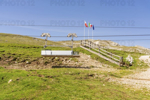 Chairlift in front of mountain landscape with flags and green slope under clear blue sky, Alpe di Siusi, Dolomites, South Tyrol, Italy