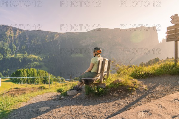 Person with bicycle helmet on a bench on a hiking trail with a view of imposing mountains, Alpe di Siusi, Dolomites, South Tyrol, Italy