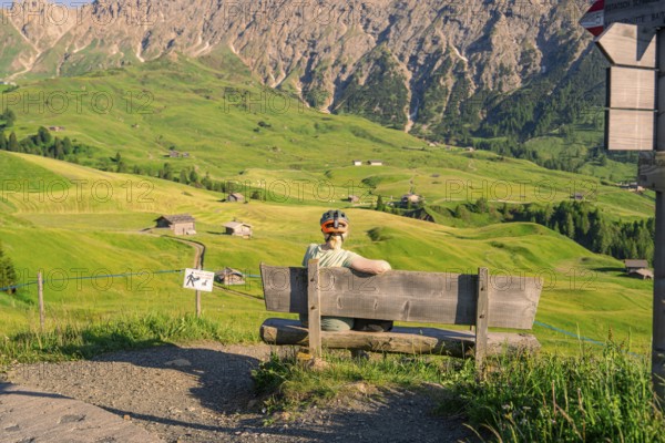 A person sits on a bench and looks out over an expansive mountain landscape with rolling hills, Alpe di Siusi, Dolomites, South Tyrol, Italy