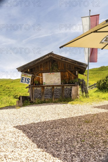 Wooden hut as an ice cream stand in a meadow in the mountains in the sunshine with flags and parasol, Alpe di Siusi, Dolomites, South Tyrol, Italy