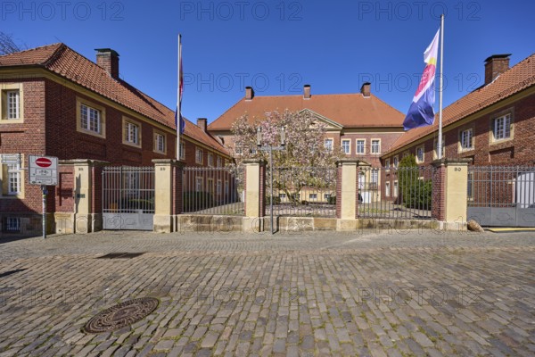 Cathedral administration, brick building, tree, cobblestone pavement, flags on flagpoles, blue cloudless sky, cathedral square, Münster, Münsterland, independent city, North Rhine-Westphalia, Germany