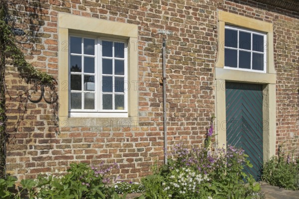 A rustic brick wall with two windows, wooden door and flowering plants, Haus Welbergen, Ochtrup, Welbergen, Münsterland, North Rhine-Westphalia, Germany