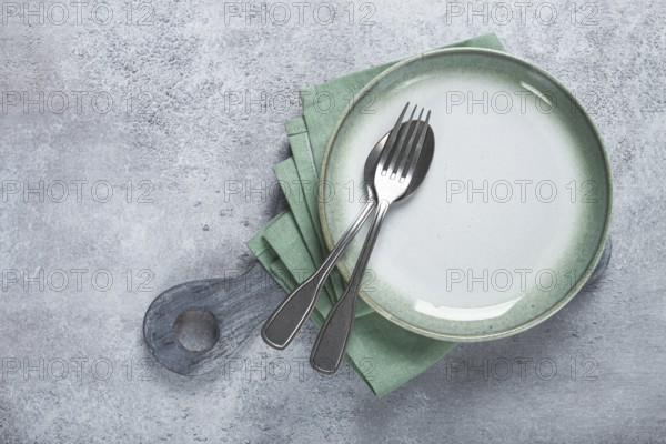 An empty plate and cutlery laid out, on a cutting board, with a green napkin, a gray table, top view