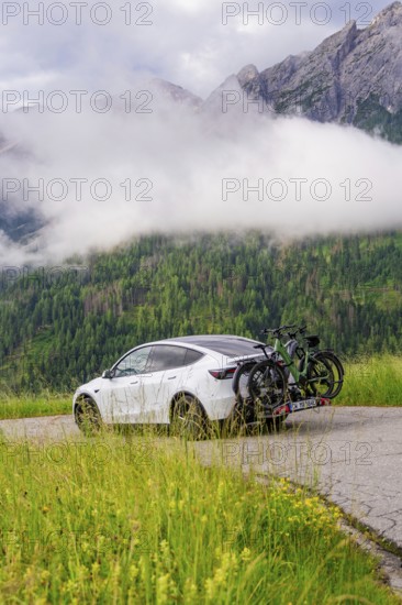 White car with bicycles on road, surrounded by misty mountain landscape and green forest, electric car on holiday, Dolomites, South Tyrol, Dolomites, Italy