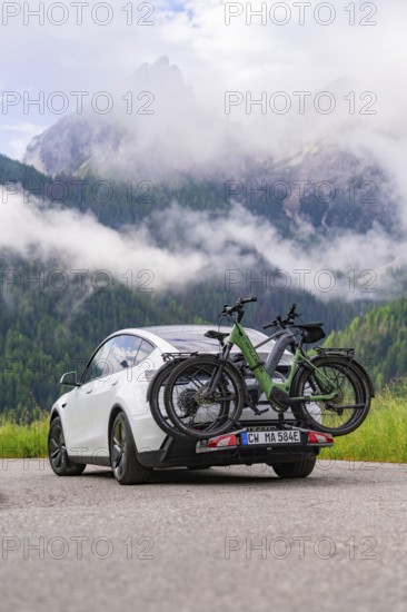 A car with bicycles drives along a road against a dramatic mountain backdrop and clouds, electric car on holiday, Dolomites, South Tyrol, Dolomites, Italy