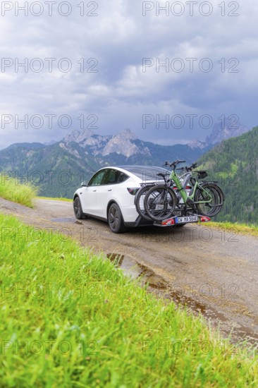 White car with bicycles on mountainous road, surrounded by green landscape and cloudy sky, electric car on holiday, Dolomites, South Tyrol, Dolomites, Italy