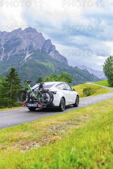 White car with bicycles on a road in a mountainous, green landscape with cloudy sky, electric car on holiday, Dolomites, South Tyrol, Dolomites, Italy