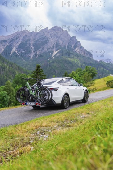 White car with bicycles on road in green, mountainous surroundings under cloudy sky, electric car on holiday, Dolomites, South Tyrol, Dolomites, Italy