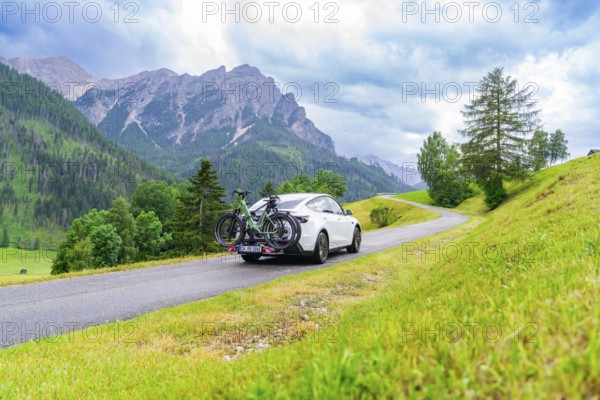 White car with bicycles on a road through green, hilly landscape with cloudy sky, electric car on holiday, Dolomites, South Tyrol, Dolomites, Italy