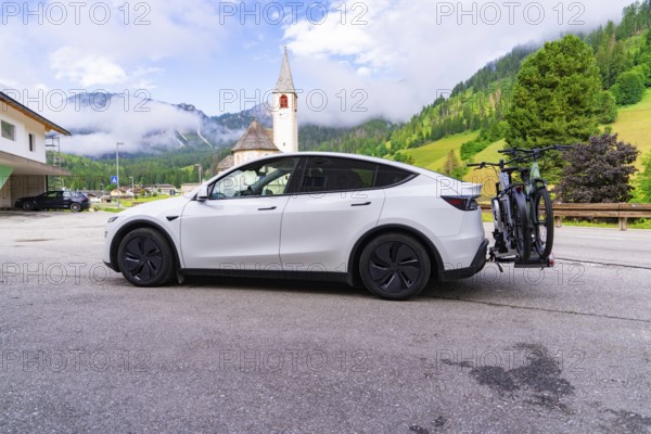 On a village road with a church in the background, a car in a mountain landscape, electric car on holiday, Dolomites, South Tyrol, Dolomites, Italy